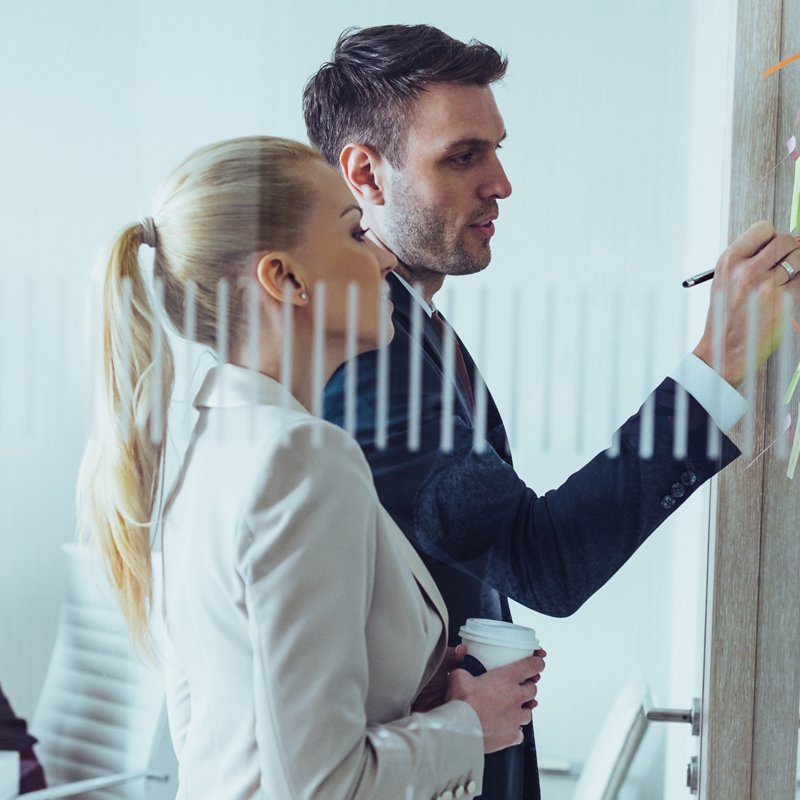 A man holding a pen writing on a board while trying to explain to a woman who stairs at what he shows her